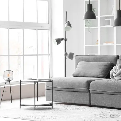 Interior of light living room with cozy grey sofa and coffee table near big window